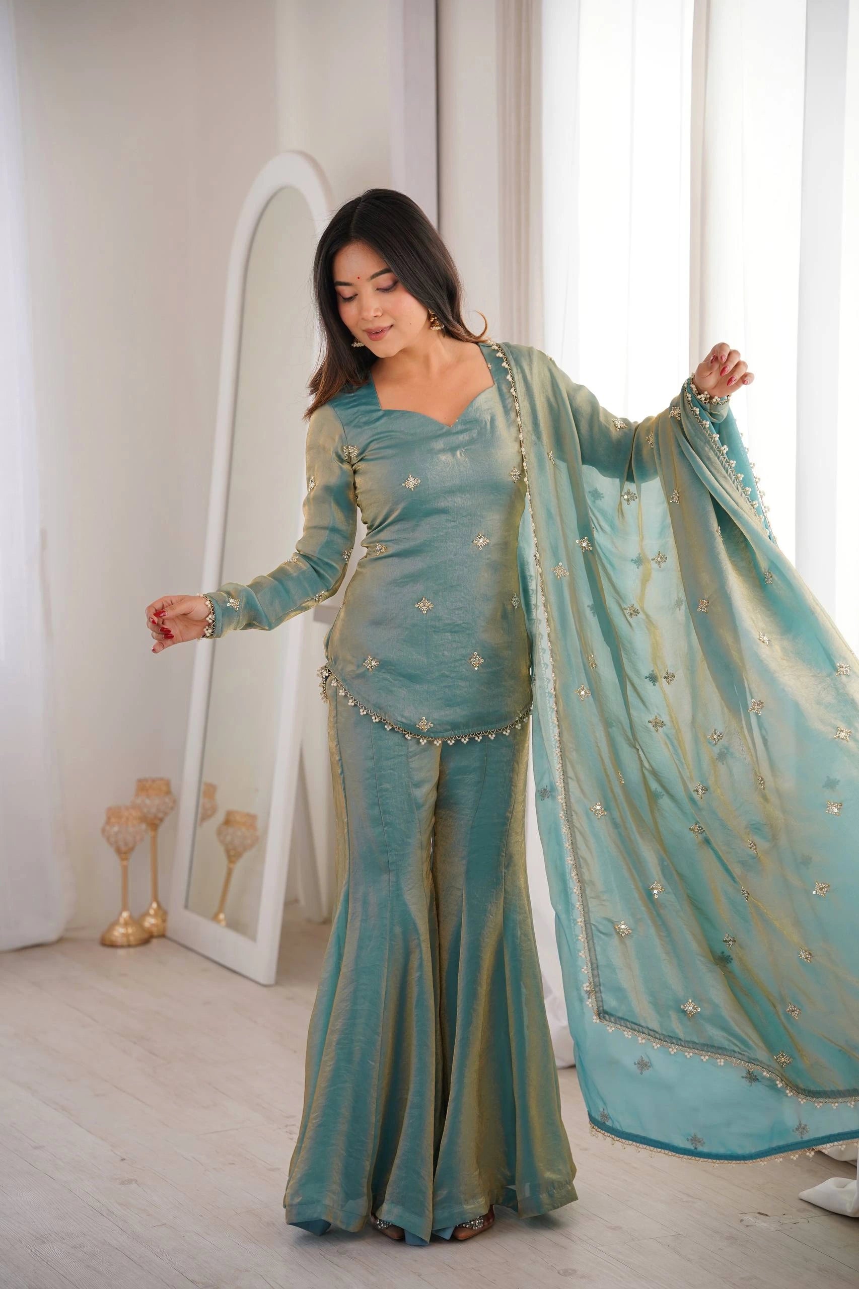 Woman in a teal traditional outfit standing in a bright room with white walls and a mirror.